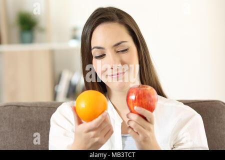 Fille de décider entre deux fruits une pomme et une orange assis sur un canapé dans la salle de séjour à la maison Banque D'Images