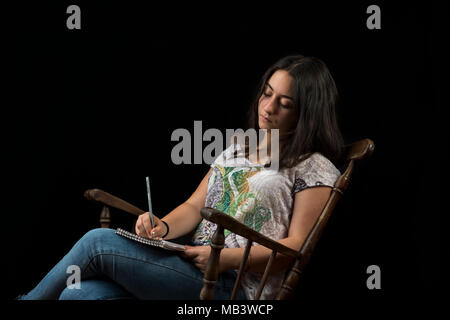 Teenage girl écrit avec un crayon dans un rocking-chair, sur fond noir Banque D'Images