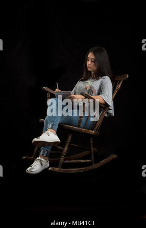 Teenage girl écrire ou dessiner avec un crayon dans un rocking-chair, sur fond noir Banque D'Images