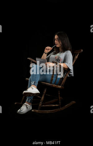 Teenage girl écrire ou dessiner avec un crayon dans un rocking-chair, sur fond noir Banque D'Images