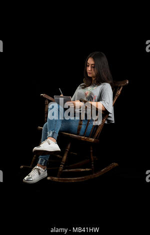 Teenage girl écrire ou dessiner avec un crayon dans un rocking-chair, sur fond noir Banque D'Images