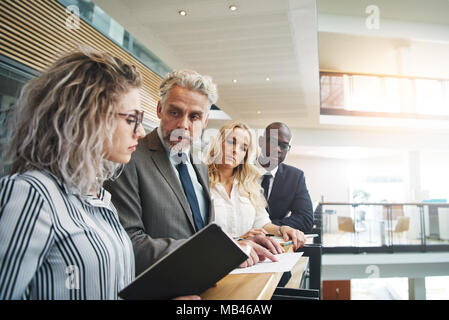 Groupe diversifié de l'accent des dirigeants discussing paperwork ensemble tout en se tenant à la rambarde dans un couloir d'un immeuble de bureaux modernes Banque D'Images