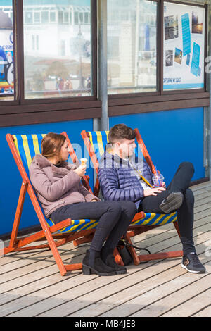 Bournemouth, Dorset, UK. 7 avril 2018. Météo France : après un début de journée humide le soleil essaie de sortir et en dépit d'être sur le côté froid de la tête aux visiteurs à la plage de Bournemouth pour profiter de la mer pendant les vacances de Pâques. Jeune couple profitez de glaces transats sur la pier Banque D'Images
