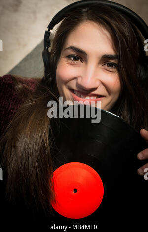 Close up of young smiling woman with headphones listening music looking at camera with vinyl records label rouge copy space Banque D'Images