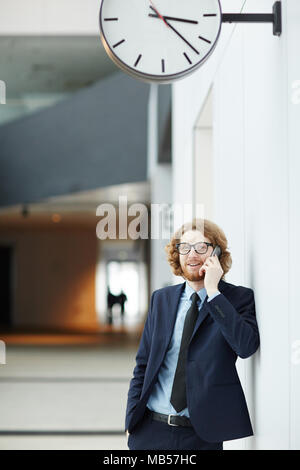 Manager en costume et barbu lunettes leaning against wall avec réveil au-dessus tout en parlant à collègue smartphone par Banque D'Images