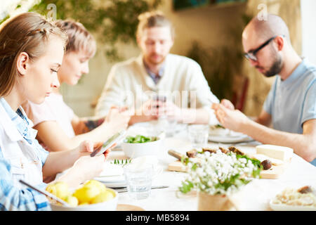 Groupe de jeunes de rechercher ou de textos en leurs gadgets en étant assis par table servi par le dîner Banque D'Images