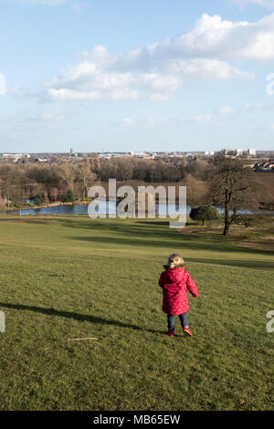Un enfant vêtu d'un manteau rouge se dresse au sommet d'une colline herbeuse, contemplant les arbres, un lac tranquille et une ville lointaine sous un ciel bleu. Banque D'Images