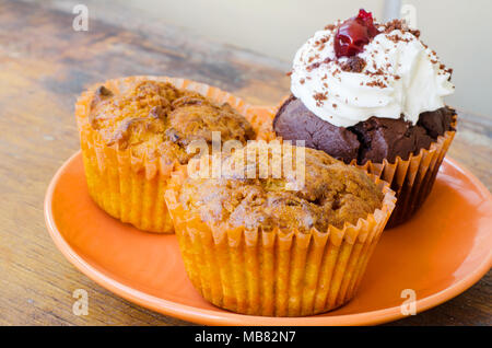 Plaque orange avec deux muffins citron et un muffin au chocolat avec crème fouettée et cerise sur une vieille table en bois Banque D'Images