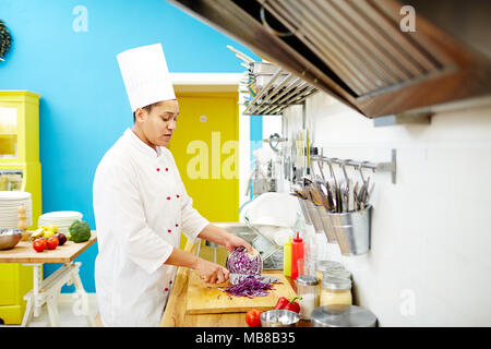 Jeune chef chopping chou violet frais tout en se tenant par le tableau pour préparer la nourriture dans la cuisine Banque D'Images
