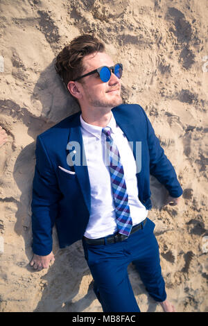 Jeune homme drôle en costume élégant se trouve sur la plage. Guy élégant est le soleil en journée ensoleillée sur le sable. Gestionnaire en lunettes inhabituelle heureux Banque D'Images