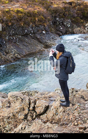 Jeune Femme prenant une photo avec un téléphone mobile debout sur des pierres à bassins, rivière fée fragile, Isle of Skye, Scotland, UK en Mars Banque D'Images