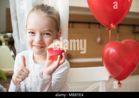 Cute girl bambin célébrer 6e anniversaire. Girl eating her birthday cupcake dans la cuisine, entouré par des ballons, showing Thumbs up. Banque D'Images
