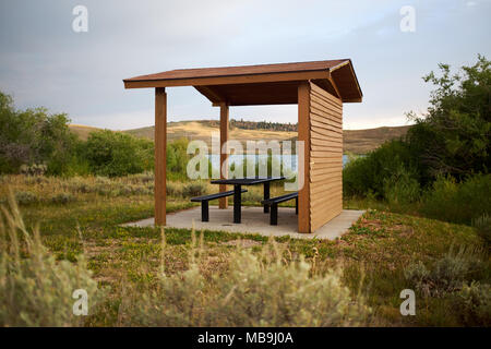 Pique-nique couverte en bois recto verso ouvert hut avec table et chaises donnant sur un lac ou une rivière dans une campagne vallonnée Banque D'Images