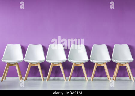 Vue de face d'une rangée de simple, moderne, blanc, bois, chaises vides contre mur pourpre dans un intérieur de style minimal Banque D'Images