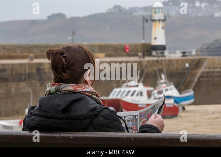 Une dame ou une femme assise sur un banc ou un siège par le côté du port à St Ives en Cornouailles lire un journal avec le mur du port derrière. Station Banque D'Images