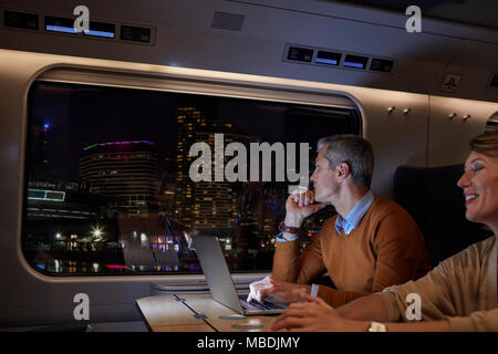 Businessman working at laptop sur train de voyageurs à la nuit, à la fenêtre en passant en ville Banque D'Images