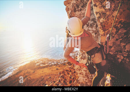 Female rock climber pour atteindre au-dessus de l'océan ensoleillé Banque D'Images