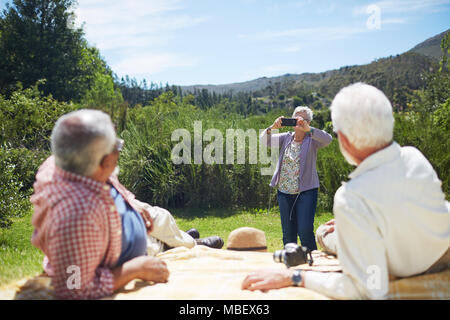Active senior woman with camera phone photographier hommes détente sur couverture de pique-nique d'été ensoleillé Banque D'Images