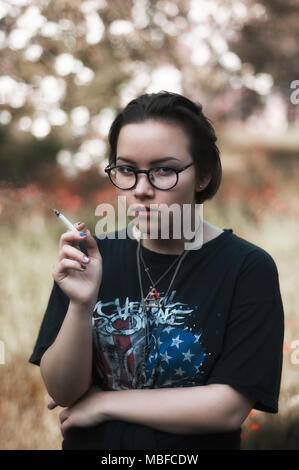 Une fille avec de courts cheveux foncés, lunettes, My Chemical Romance un t-shirt et d'une cigarette dans un parc. Banque D'Images