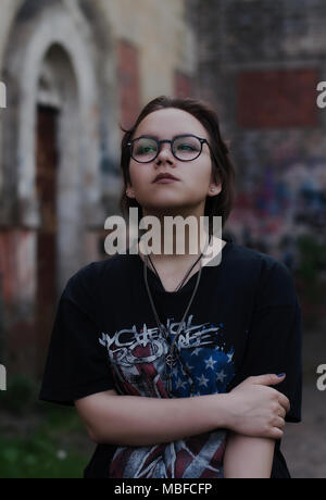 Portrait d'une d'une fille avec des cheveux noirs courts, lunettes, My Chemical Romance un t-shirt et colliers debout à côté d'un vieux bâtiment avec des graffitis. Banque D'Images