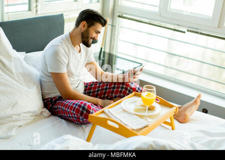 Jeune homme de manger le petit déjeuner au lit pendant l'utilisation de téléphone mobile Banque D'Images