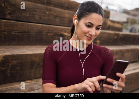 Pretty smiling brunette avec téléphone sur escaliers vêtu de blanc écouteurs, une chemise marron et les cheveux attachés en arrière Banque D'Images