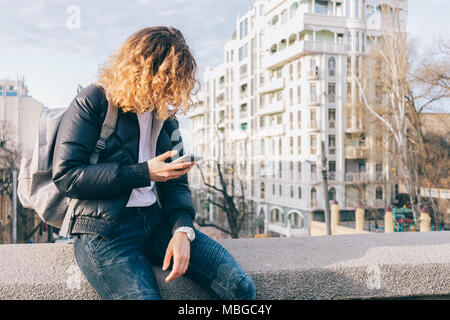 Young woman texting on smart phone dans la ville. Jeune fille rousse frisée est assis sur le parapet du pont au jour de printemps ensoleillé. Banque D'Images