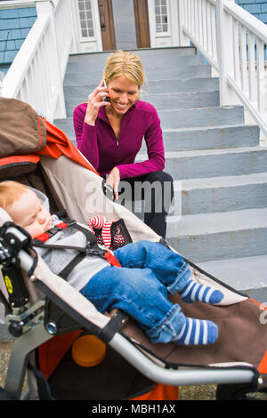 Une mère se trouve sur l'avant des escaliers en conversation sur son téléphone portable tout en un petit garçon dort dans une poussette. Banque D'Images