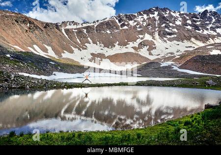 Randonneur dans une chemise orange sautant avec prends part près du lac dans les montagnes enneigées Banque D'Images