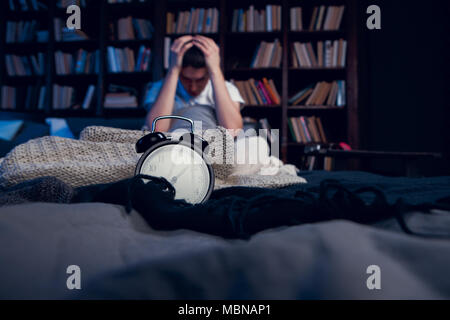 Photo de brunet à l'insomnie assis au lit avec réveil Banque D'Images