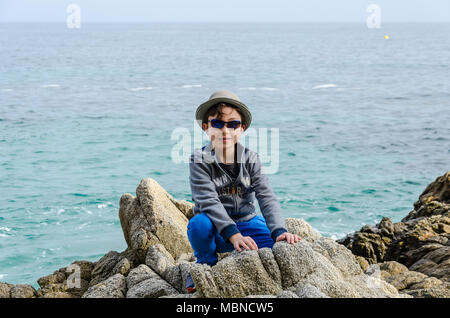 Un jeune garçon s'accroupit vers le bas sur des rochers près de la mer et de pose pour une photo. Banque D'Images