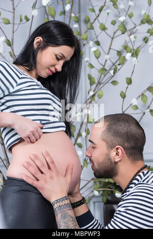 Beau couple international dans des pulls à rayures se regardent, musulman, homme assis et épouse sa femme enceinte dans la chambre à coucher Banque D'Images