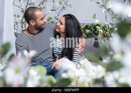 Voir à travers les fleurs sur charming international dans deux pulls à rayures regarder les uns les autres et s'étreindre tout en étant assis sur le lit dans la chambre à coucher Banque D'Images