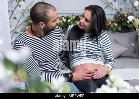 Heureux couple international chez chandails, muslim man hugging ventre de sa femme enceinte, assise sur le lit dans la chambre à coucher Banque D'Images