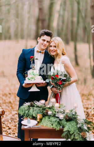 Beautiful wedding couple posant avec un gâteau dans les mains près de la table vintage. Forêt d'automne background Banque D'Images