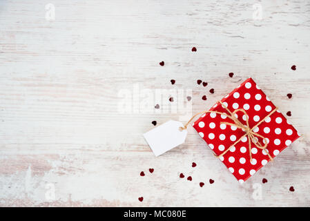 Vue de dessus d'une boîte-cadeau en pointillés rouges sur fond de bois blanc. Copier l'espace. Note vide. Sequins en forme de coeur dispersés. Banque D'Images