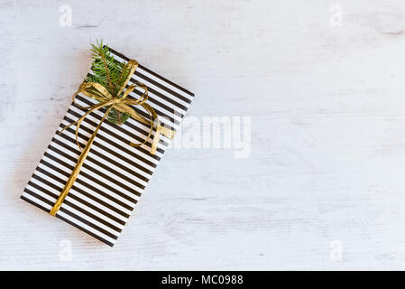 Boîte-cadeau enveloppé dans du papier à rayures noir et blanc avec du ruban d'or et de branches de sapin blanc sur un fond de bois. Concept de Noël. Banque D'Images