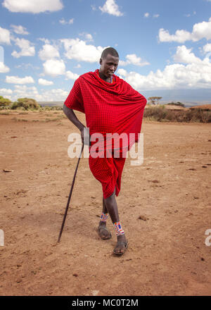 Pas de village près d'Amboselli park, Kenya - 02 avril, 2015 : Inconnu guerrier Masai qui pose pour les touristes en robe rouge lumineux traditionnel, s'appuyant sur son Banque D'Images