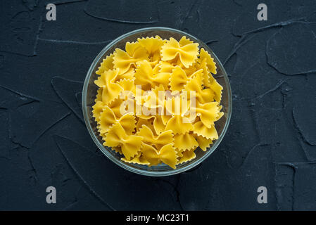 Macaroni farfalle pâtes dans un bol en verre sur un fond texturé noir, au centre close-up par le haut. Banque D'Images