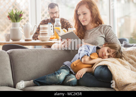 Fils dormir couché sur les genoux de sa mère alors qu'elle est en train de lire un livre et père de manger dans l'arrière-plan Banque D'Images