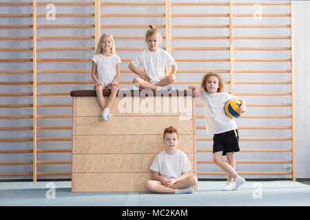 Garçon avec une balle à côté d'un saut en bois boîte sur laquelle smiling enfants sont assis dans le hall de sport Banque D'Images