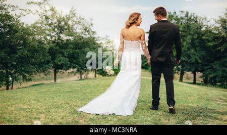 Vue arrière de la belle jeune femme dans une robe de mariage qui tient la main de l'époux et la marche dans le parc. Couple holding hands and walking outdoors sur Banque D'Images