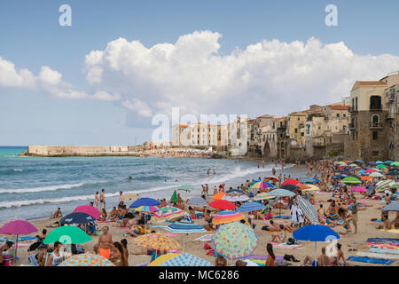Cefalu, Sicile, Italie, des foules de gens sur la plage de sable de jouir de leurs vacances sur une journée ensoleillée d'août avec la vieille ville bâtiments comme un ta Banque D'Images