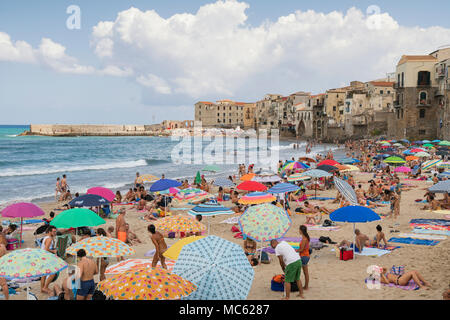 Cefalu, Sicile, Italie, des foules de gens sur la plage de sable de jouir de leurs vacances sur une journée ensoleillée d'août avec la vieille ville bâtiments comme un ta Banque D'Images