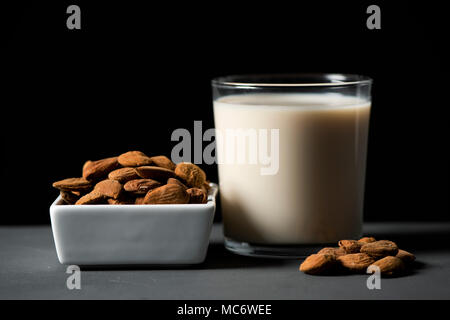 Libre d'un bol en céramique blanche avec des amandes et un verre de lait d'amande sur une table rustique en bois gris, sur un fond noir Banque D'Images