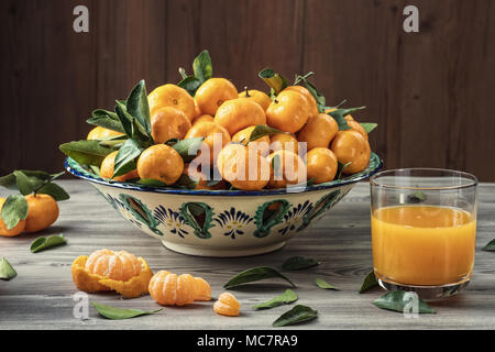 Tangerines fraîches (mandarines) en plat de céramique, verre de jus sur la table en bois gris et marron plank arrière-plan. La lumière du jour. Concept d'aliments sains. Banque D'Images