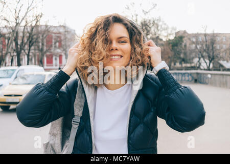 Jeune femme avec des cheveux bruns bouclés met sur le capot en marchant. Mignon femelle traveler s'amusant à jour de printemps dans la ville. Banque D'Images