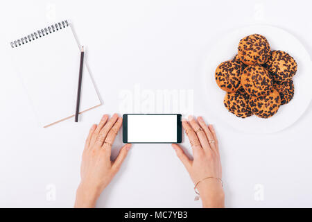 Vue de dessus de femmes portez des bagues et bracelet holding smart phone avec écran vide près de cookie et blank notepad avec crayon sur blanc backgroun Banque D'Images