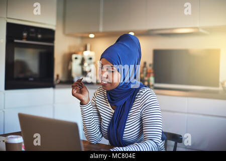 Souriante jeune femme entrepreneur arabe portant un hijab assis à sa table de cuisine à l'aide d'un ordinateur portable tout en travaillant de chez vous. Banque D'Images