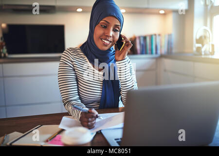 Jeune femme entrepreneur arabe portant un hijab assis à sa table de cuisine l'utilisation d'un cellulaire et à l'aide d'un ordinateur portable tout en travaillant de chez vous. Banque D'Images